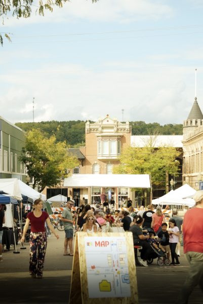 Crowds gather on a busy street and bright day, with a music stage in the background.